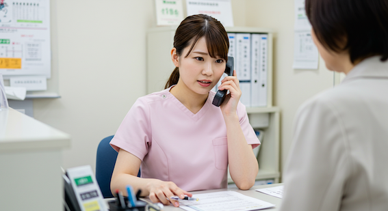 A female Japanese medical receptionist in a clinic with office dress is taking a phone call while a patient is waiting at the reception desk. She looks apologetic and is struggling with the work spread out on her desk in front of the patient.